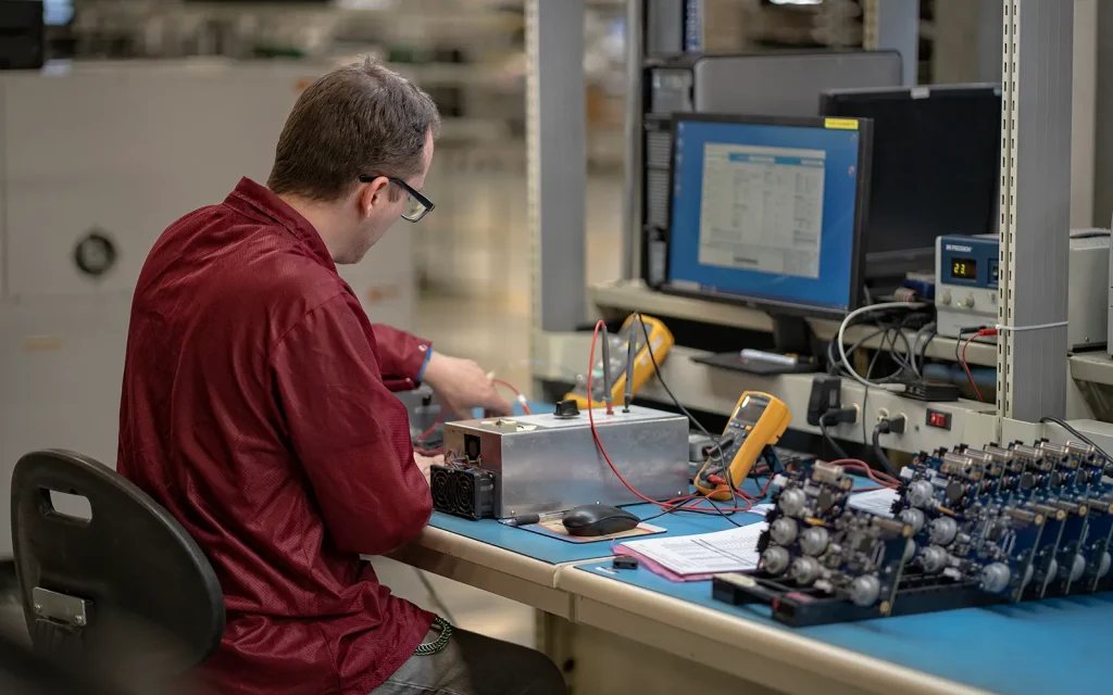 Technician at a functional test bench with a fixture, meters, and assembled boards at Intervala